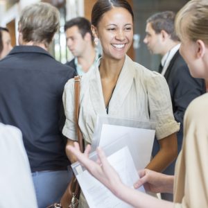 Portrait of two smiling women holding files and talking, standing in crowded lobby